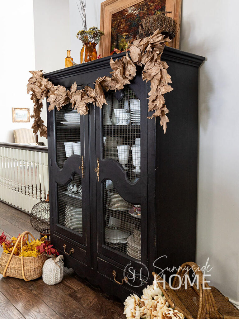 A Fall garland hung across a black painted china hutch. The garland is made from Amazon packing paper. Also styled with basket of fall leaves