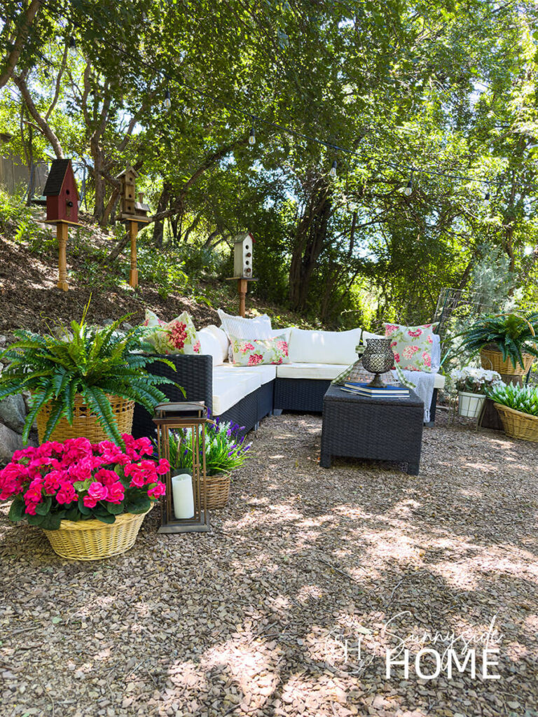 Outdoor living room with a sectional seating area styled with pillows and throw blanket, coffee table with candles and holder, assorted potted plants in containers and baskets with ferns and flowers, bird houses.