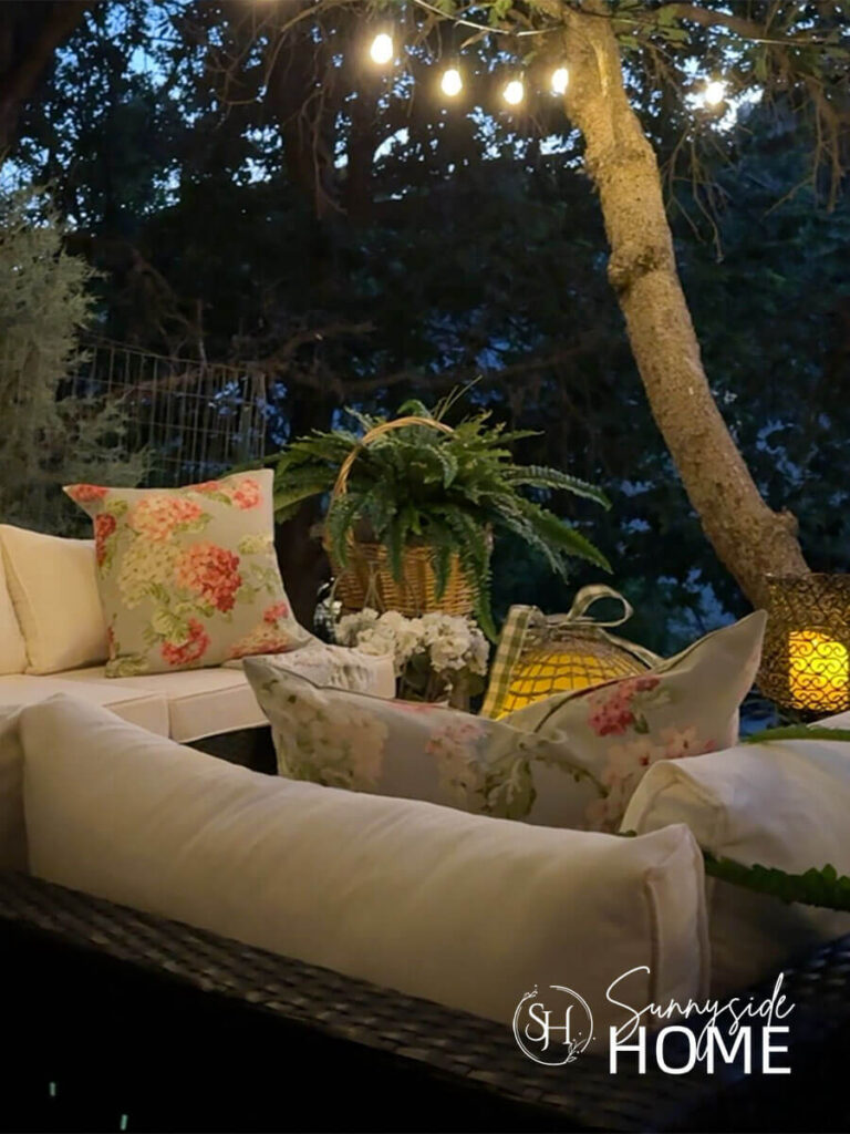 Outdoor living room in the evening with candle lit lanterns, potted plants, seating area and overhead string lights.