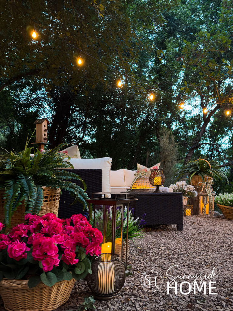 Outdoor living room in the evening with candle lit lanterns, potted plants, seating area and overhead string lights.