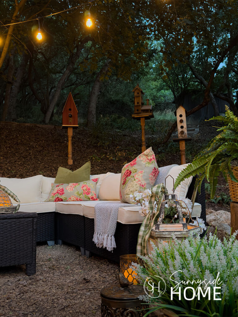 Outdoor living room in the evening with candle lit lanterns, potted plants, bird houses, seating area and overhead string lights.