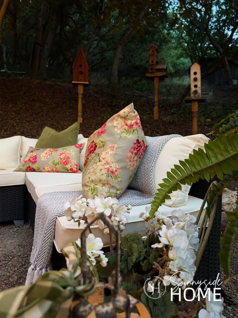 Outdoor living space with cozy floral pillows, throw blanket, potted plants and birdhouses.