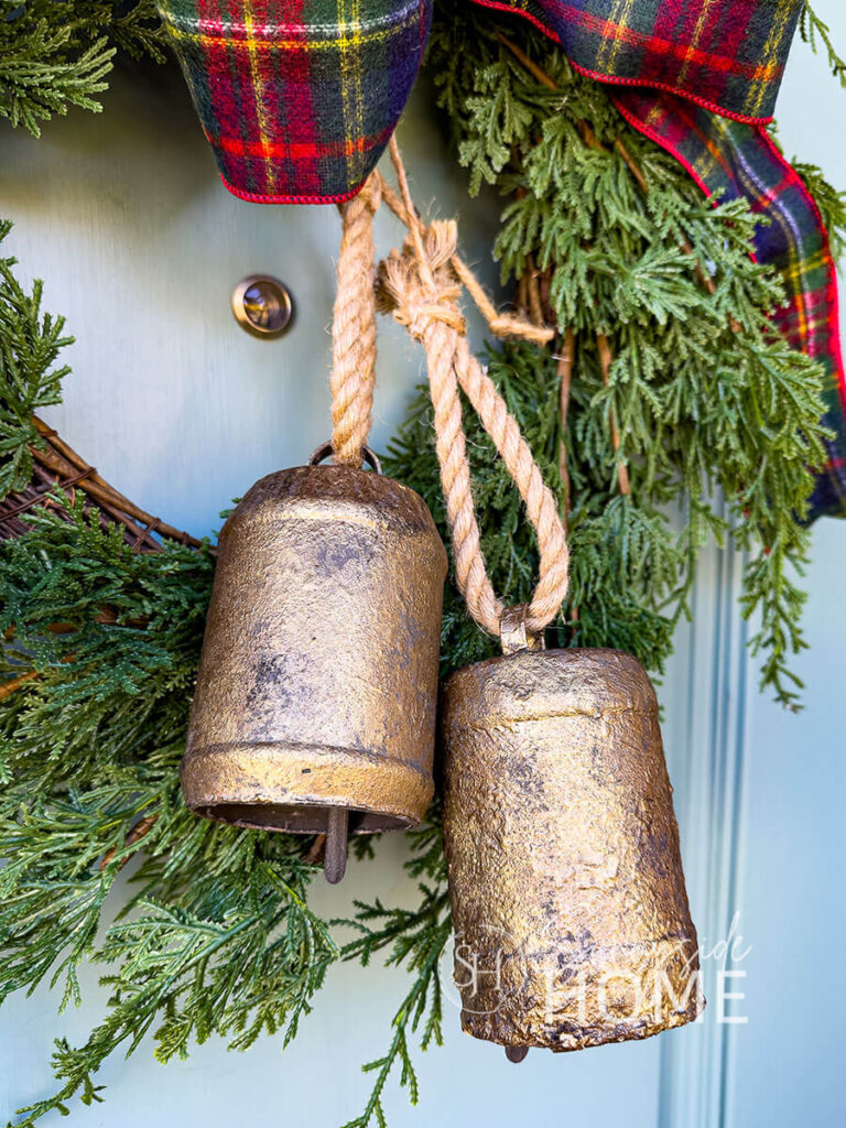 Peanut butter jars are repurposed into Christmas bells useing paint and rub and buff. Styled hanging from an evergreen wreath with a plaid bow.