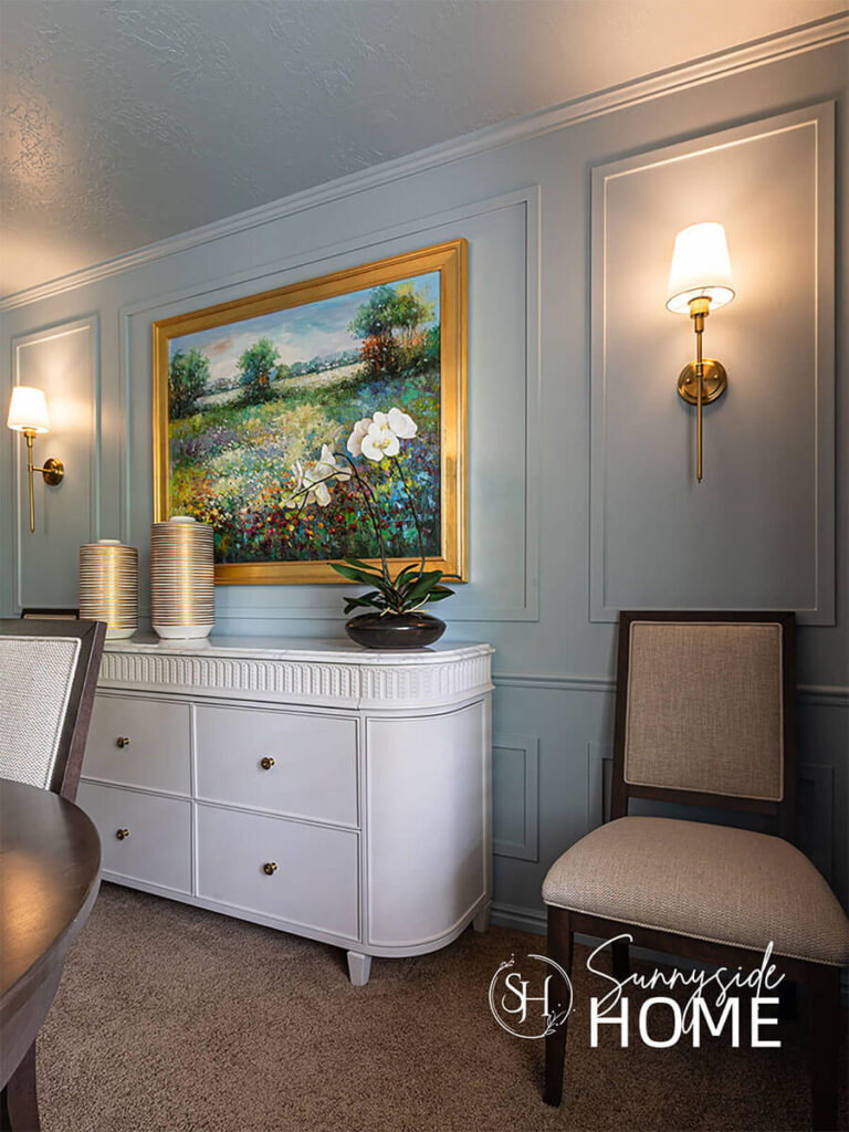 A dining room is color drench in a dusty blue and styled with a white buffet. Floral landscape art on the wall with gold sconces.