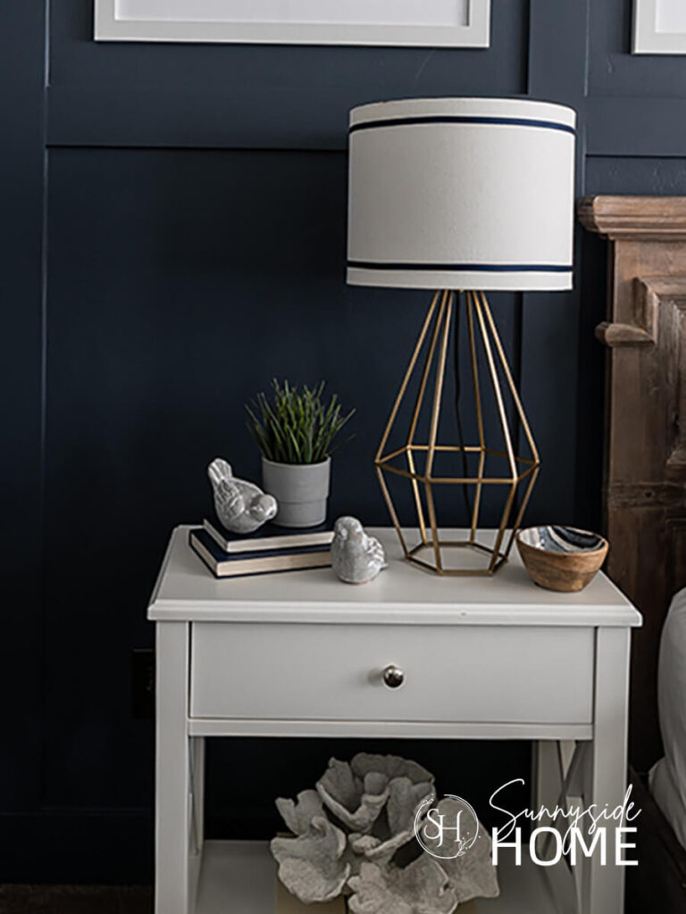 Embellished white lampshade with 2 rows or blue grosgrain ribbon on a gold geometric lamp base on a white nightstand.