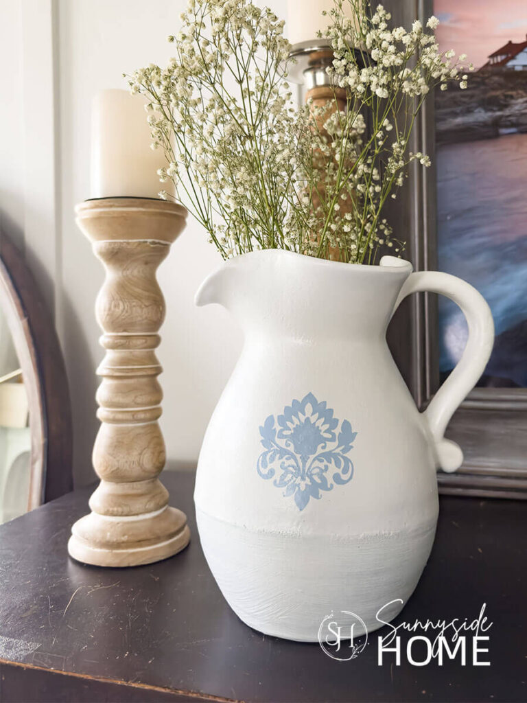 A white pitcher is stenciled with a blue damask pattern, styled with babies breath and mango wood candlesticks.