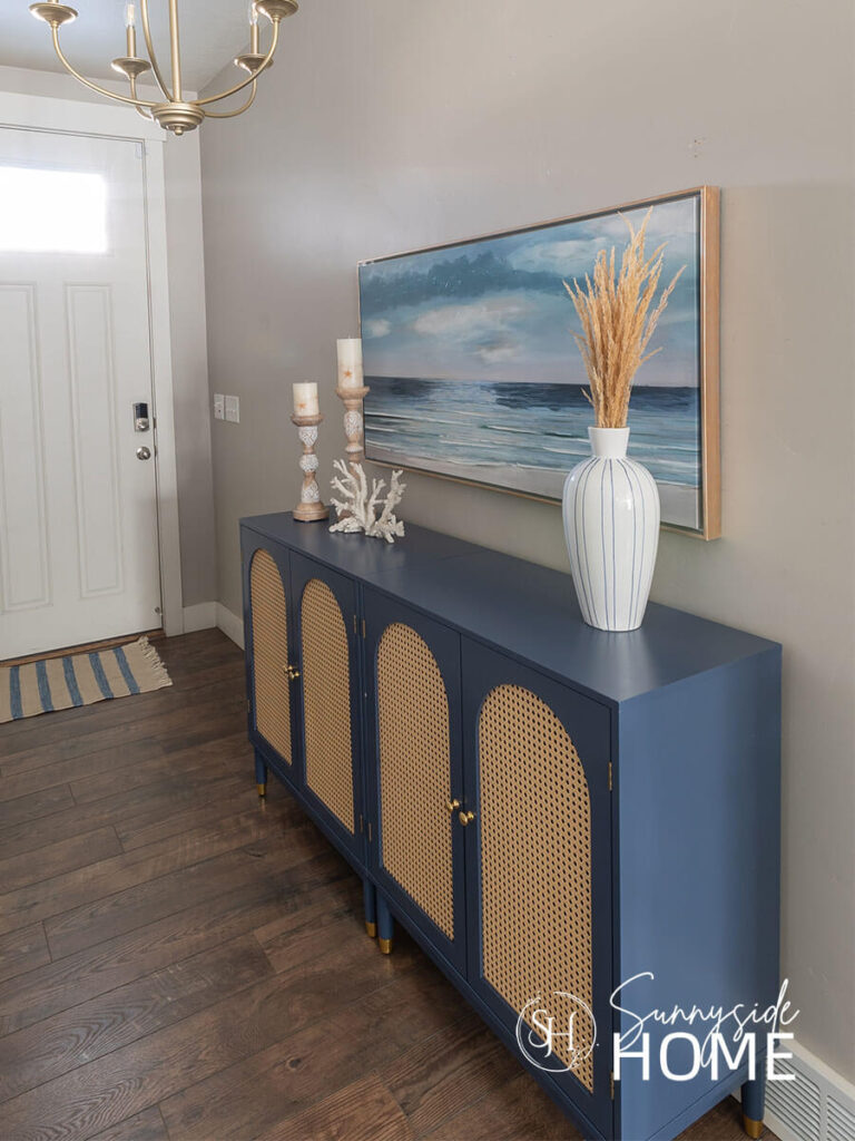A blue painted cabinet with arched rattan doors in an entryway, styled with a blue and white striped vase, white coral, a pair of mango wood candlesticks and a seascape painting.