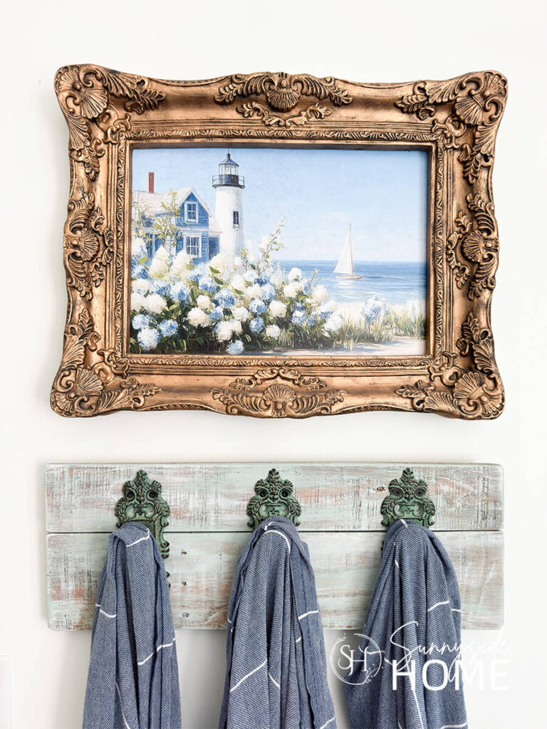 Blue and white coastal art in a gold frame styled in a bathroom above a towel rack.