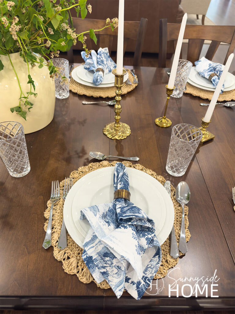 A neutral styled dining table with a jute placemat, white plates with a blue and white napkin in a brass napkin ring.