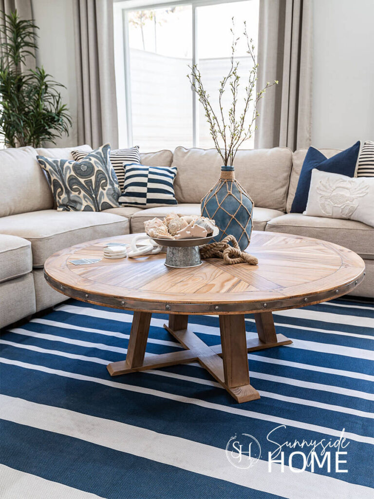 A cream color sectional is decorated blue and white pillows. A round wood coffee table styled with a blue jug vase with green branches, a tiered tray with sea shells, rope and marble coasters.