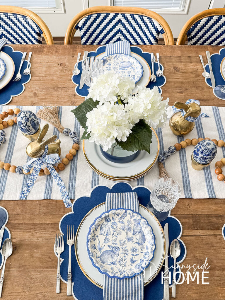 Decorating a Dining table, set with blue and white place mats, topped with white gold rimmed plates, blue and white stripe napkin and a bunny plate. For the centerpiece there is a blue color block vase with white hydrangeas on a blue and white stripe table runner