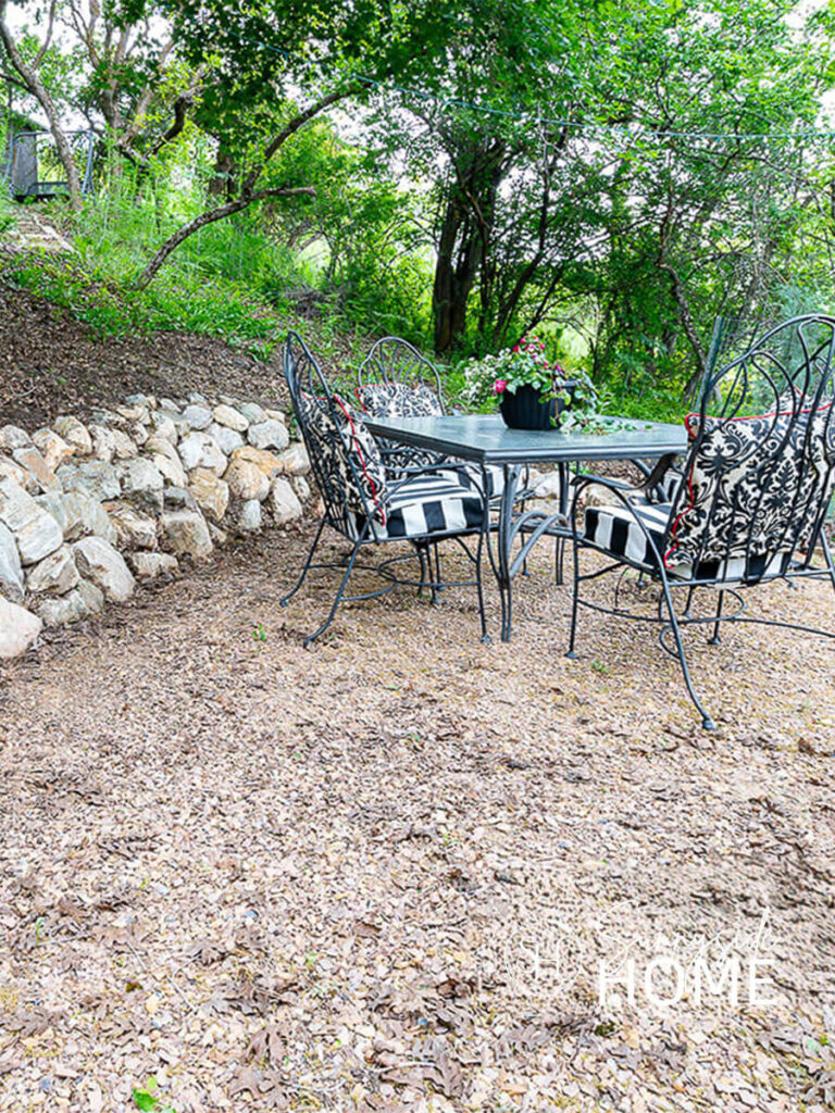 Outdoor Living Room before makeover with an old thrifted outdoor dining table with black and white cushions and a stone retaining wall.