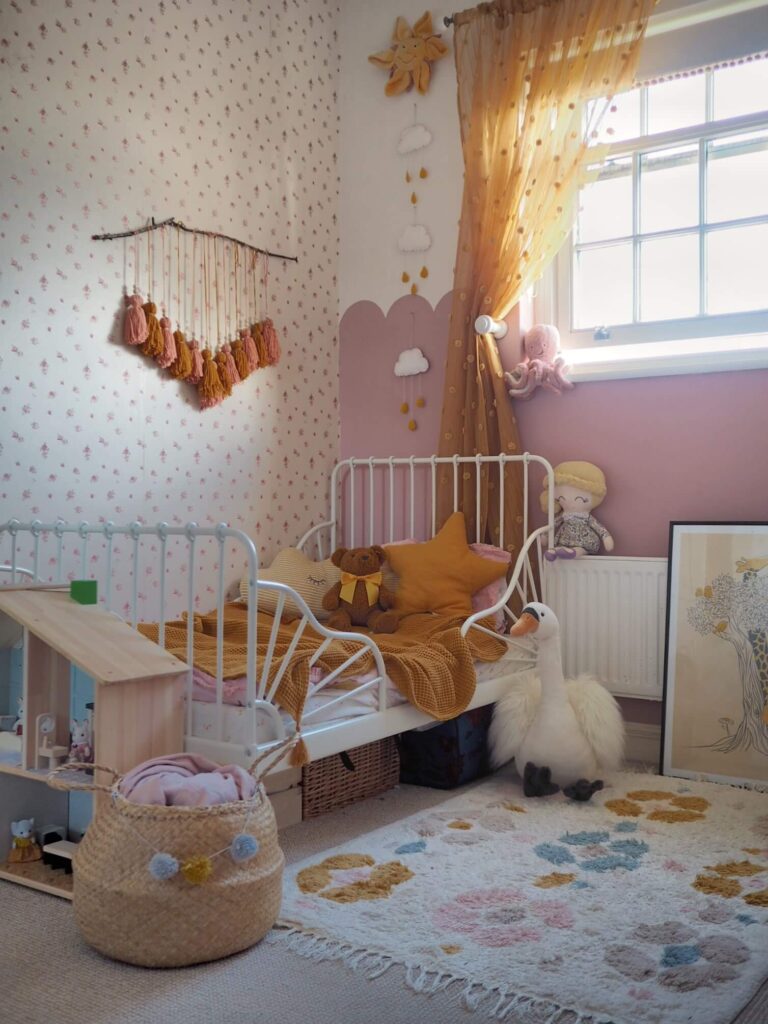 A pink painted scalloped wainscoting is painted on the wall of this little girls bedroom. Styled with yellow sheer curtains, pompom embellished shade, shite bed with yellow bedding, and child's toys.