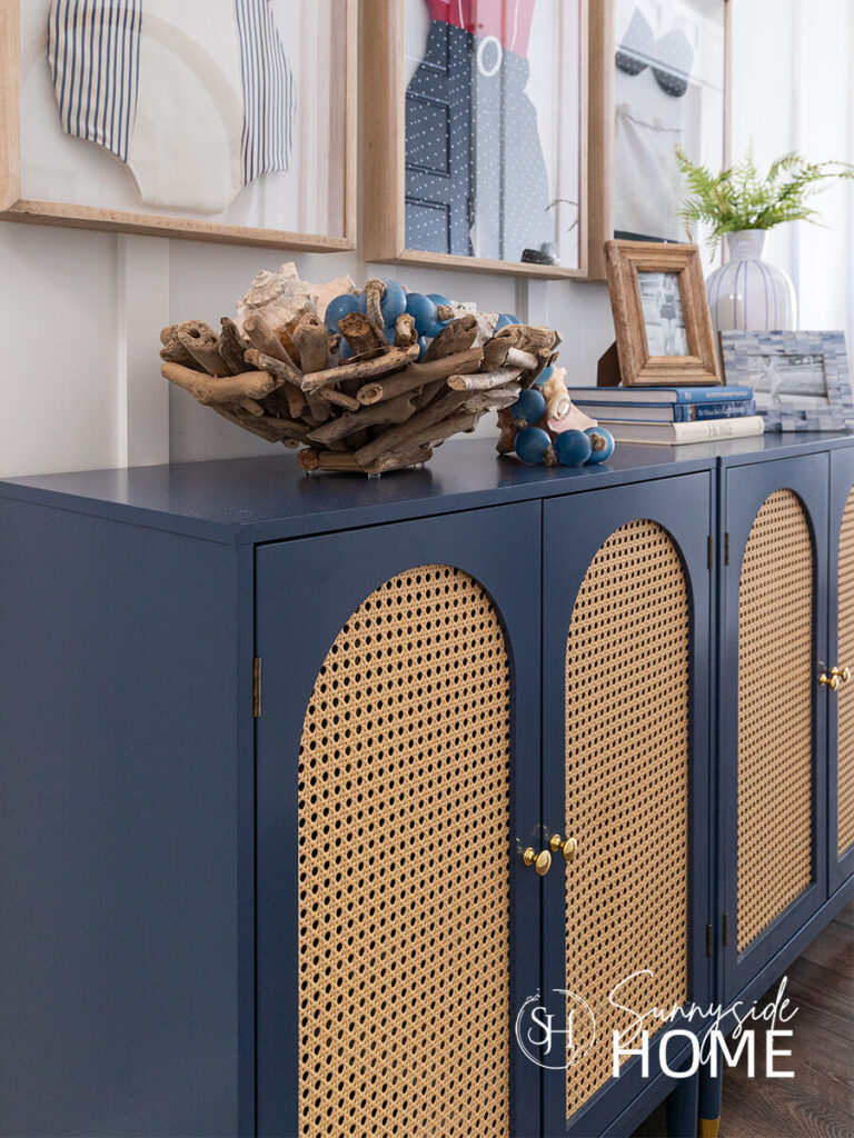 Navy blue entryway cabinet with arched rattan doors styled with a driftwood bowl filled with seashells and blue glass beads for summer.