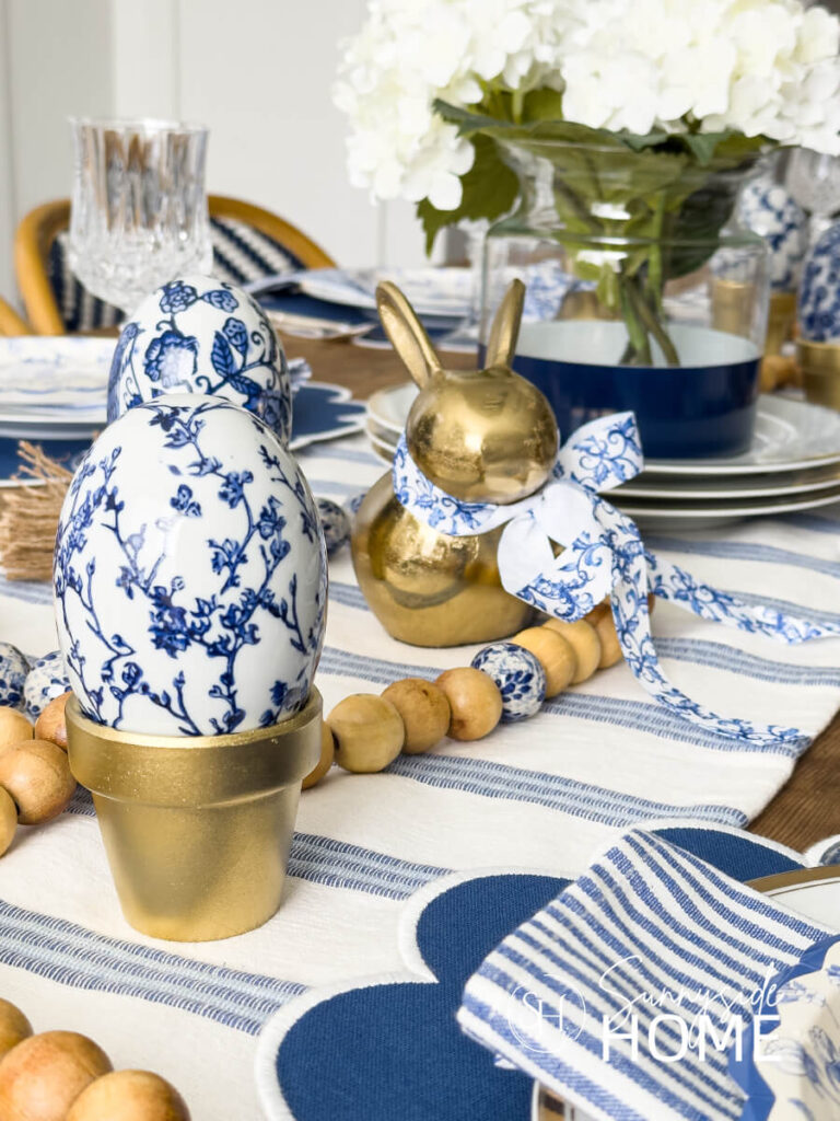 A blue and white tablescape with blue and white ceramic chinoiserie eggs in metallic gold pots. The dining table is also styled with a blue and white runner, wood bead garland with blue and white chinoiserie beads, blue and white placemats and napkins and a blue color block vase with white hydrangeas.