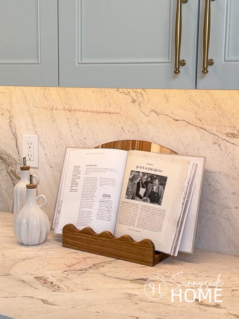 Styled in a kitchen on a stone countertop is a wood scalloped cookbook stand with white oil bottles.
