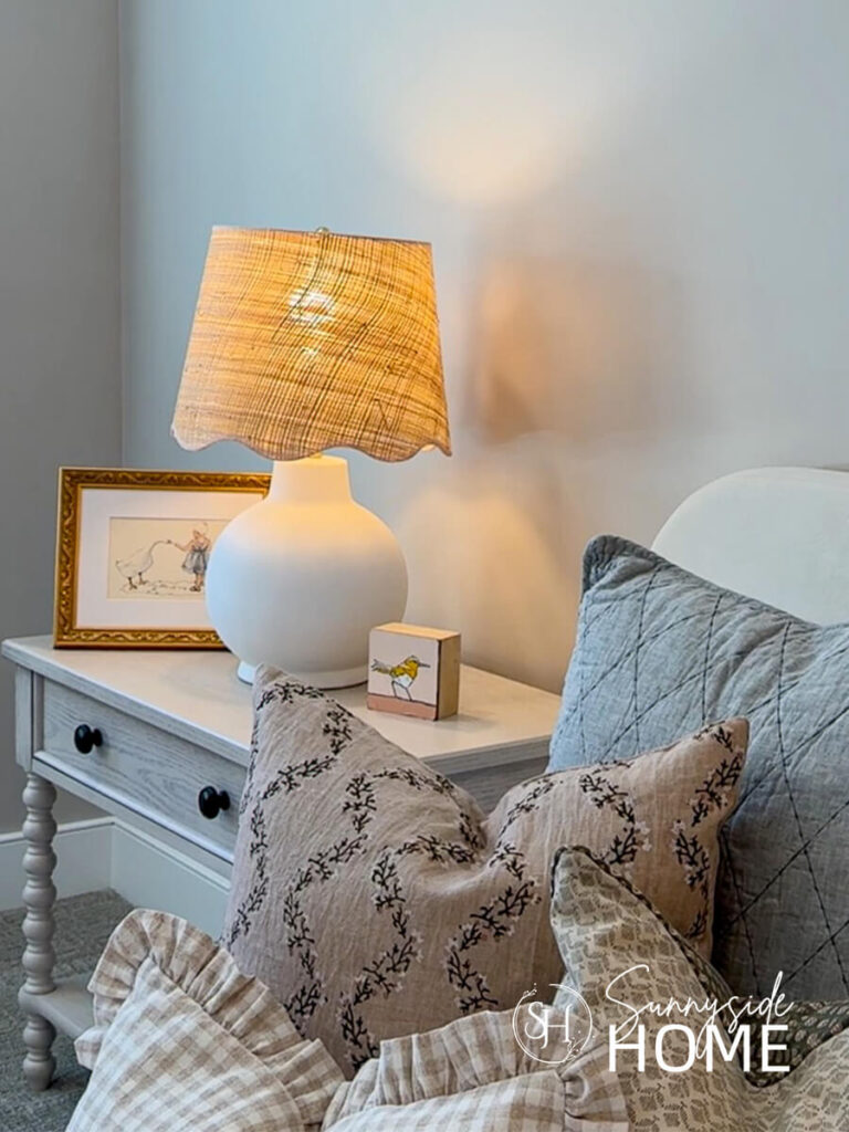 A rattan scalloped lamp shade adds texture to this feminine bedroom styled with framed art and assorted pillows.
