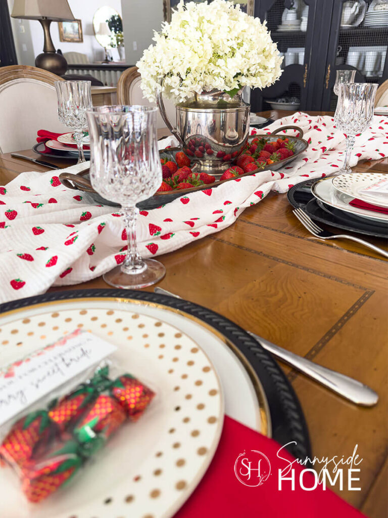 Berry in love themed tablescape for bridal shower ideas. Place setting features a black charger topped with a gold rimmed plate, red napkin, gold polka salad plate and a strawberry candy party favors. The center piece is white hydrangeas in a silver pitcher on a tray filled with fresh strawberries with a strawberry printed table runner.