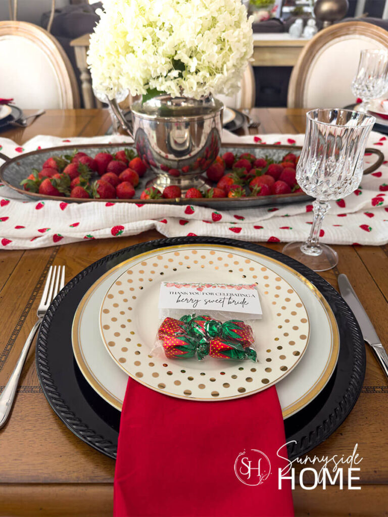Berry in love themed tablescape for bridal shower ideas. Place setting features a black charger topped with a gold rimmed plate, red napkin, gold polka salad plate and a strawberry candy party favors. The center piece is white hydrangeas in a silver pitcher on a tray filled with fresh strawberries with a strawberry printed table runner.