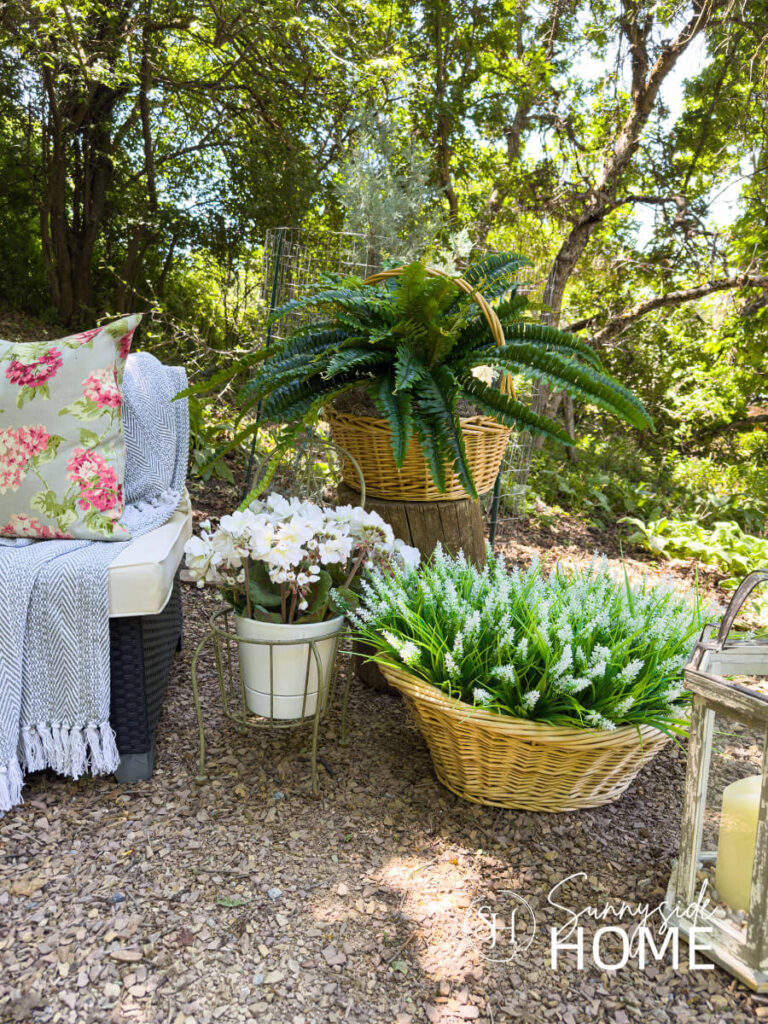 An assortment of thrift store pots and basket with potted flowers and ferns along side an outdoor sectional with throw pillow and blanket.