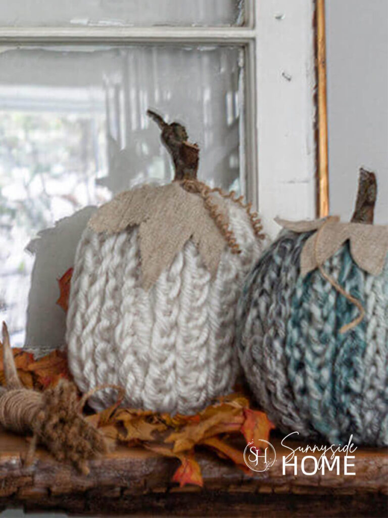 A pair of DIY fall decor finger knit pumpkins in ivory and varigated green with a twig stem and linen leaves, styled with preseved maple leaves and a vintage window.
