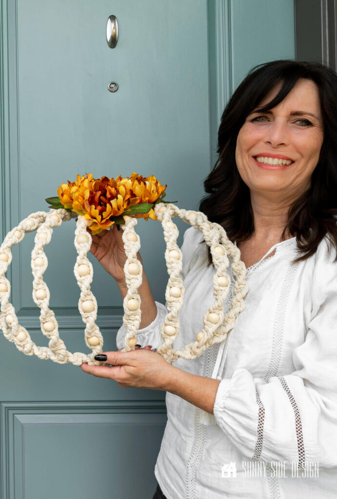 A woman holds a macrame pumpkin wreath embellished with orange peonies, in front of a blue door. The wreath is made with a basic dollar tree wire pumpkin form, cotton cording and natural beads.