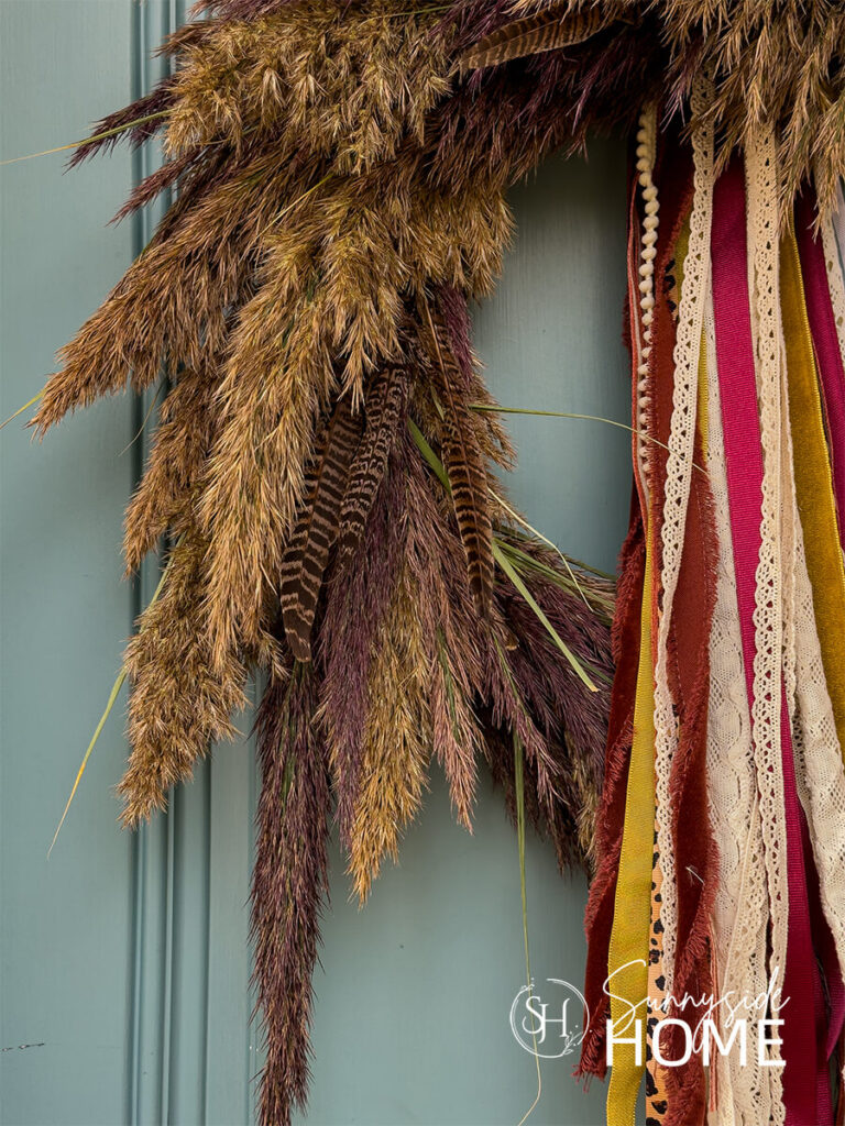 Beautiful Pampas Grass Wreath for Fall hung on a blue front door embellished with a fall ribbon bundle and pheasant feathers.