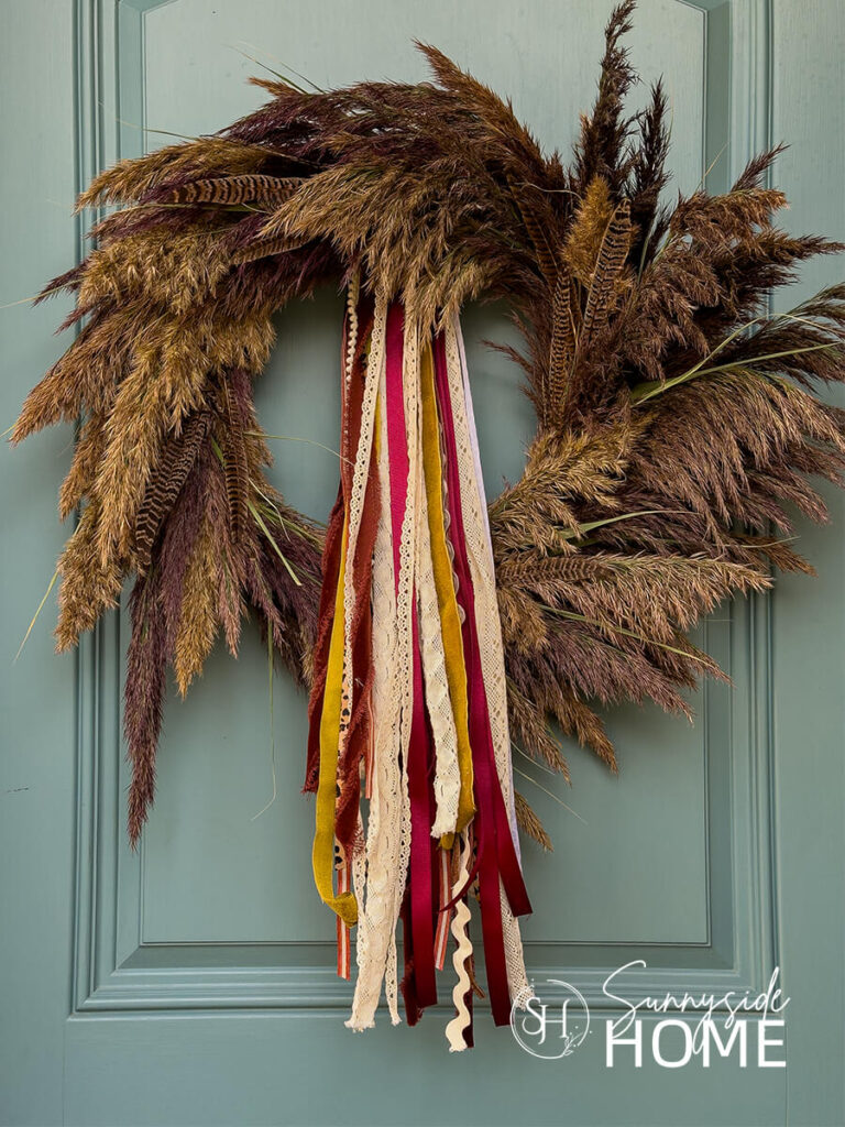 Beautiful Pampas Grass Wreath for Fall hung on a blue front door embellished with a fall ribbon bundle and pheasant feathers.