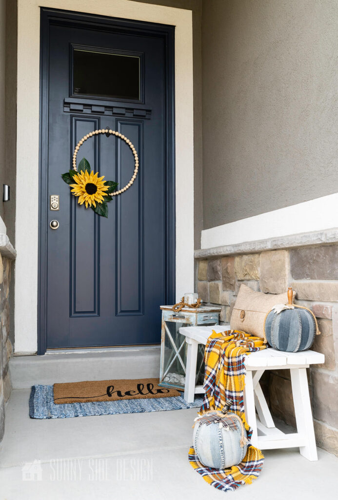 A simple modern bead wrath embellished with a giant sunflower on a navy blue door. The porch is also styled with a mustard plaid throw on a white farmhouse bench with denim pumpkins.