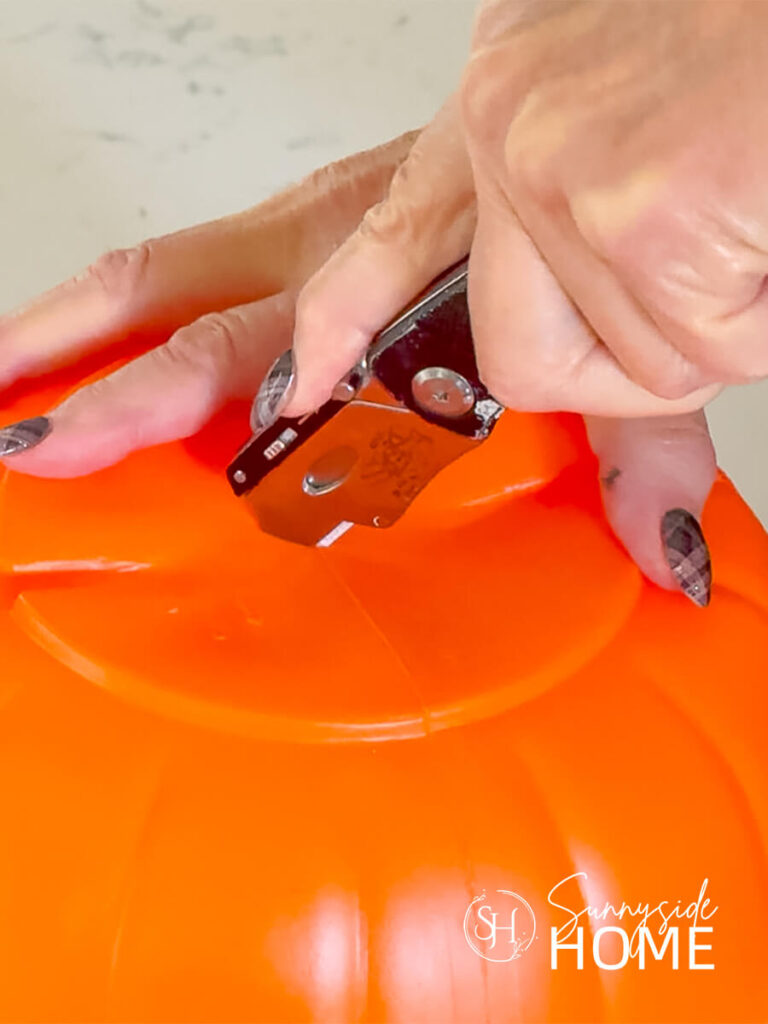 A woman uses a utility knife to cut a "X" in the bottom of an orange plastic pumpkin bucket.