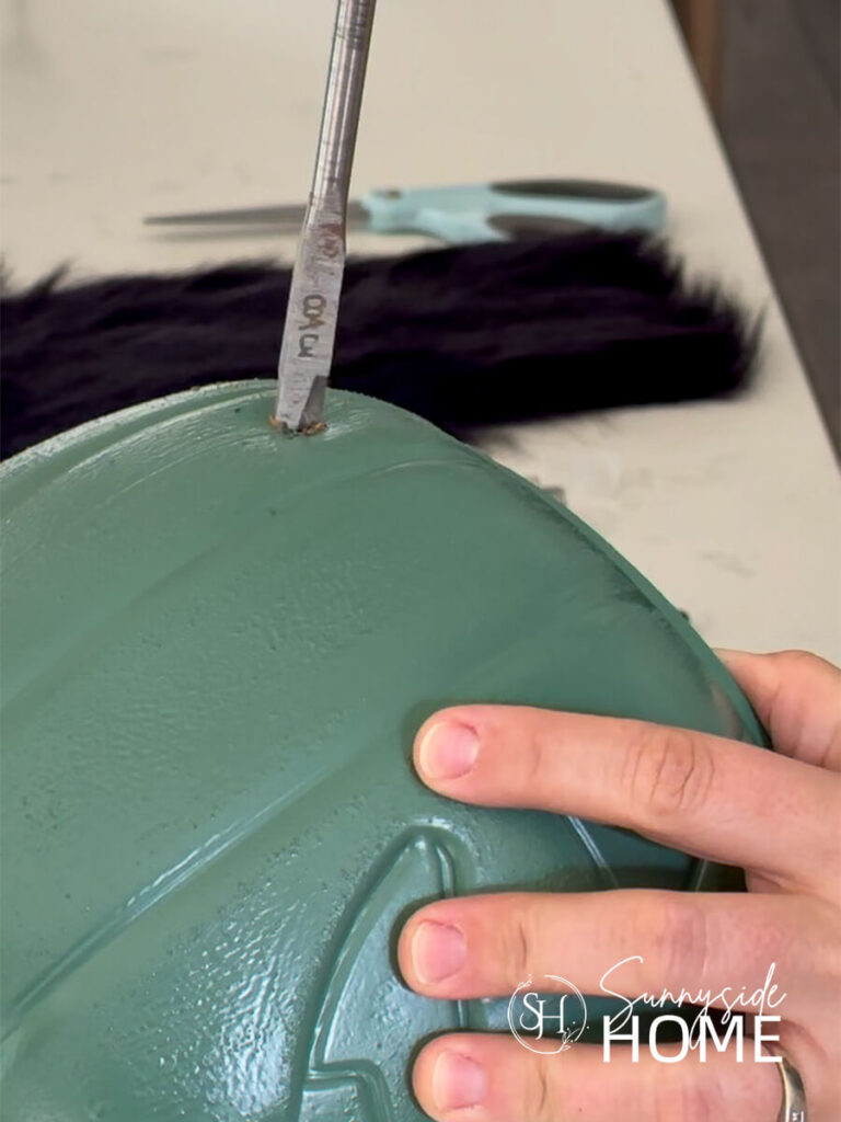 A woman drills a hole into a pumpkin bucket painted green for Halloween decor.