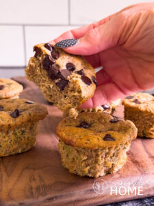 Woman holds half a protein zucchini muffin with chocolate chips over a charcuterie board with protein muffins.