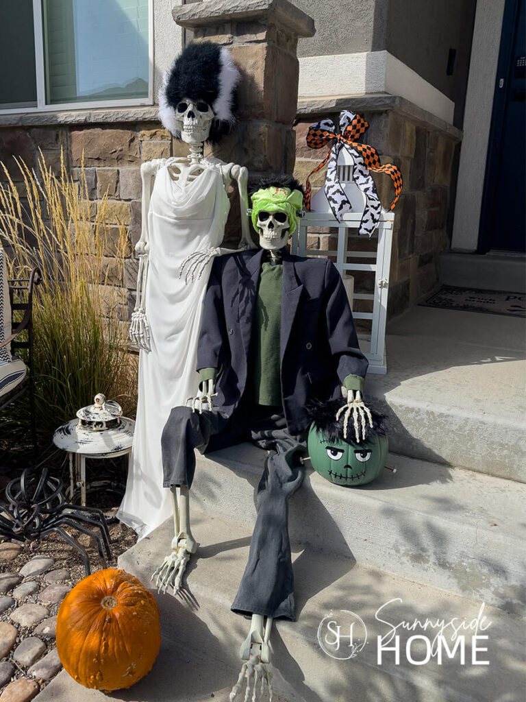 The front porch stairs styled with Halloween decor including skeletons dressed up as the bride of Frankenstein and Frankenstein and a pumpkin bucket turned into a Frankenstein head.