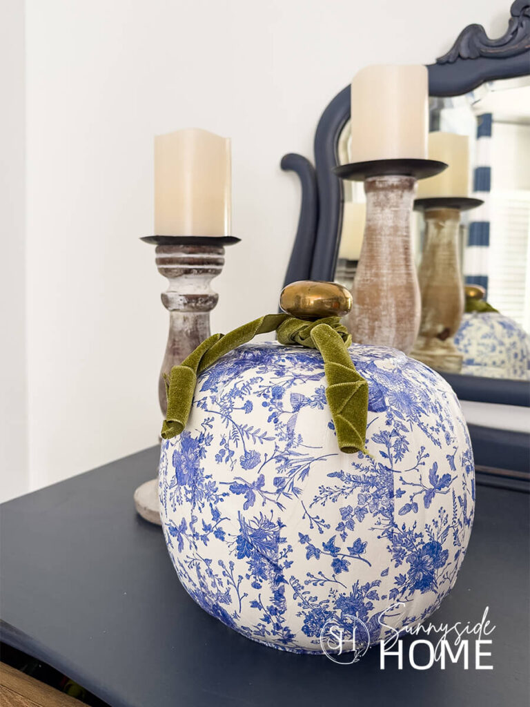 Styled blue and white chinoiserie decoupage pumpkins with pretty floral napkins, embellished with green velvet ribbon, styled on a navy blue dresser with wood candleholders.
