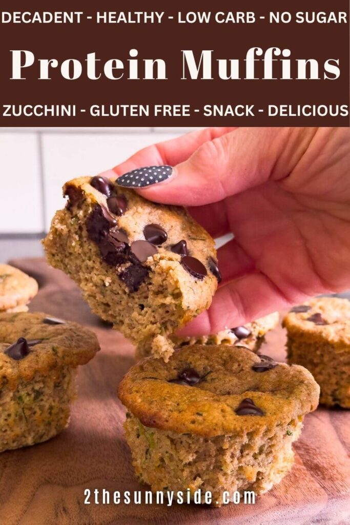 Pinterest image, woman holding half a protein zucchini muffin with creamy dark chocolate chips over a tray of healthy gluten-free high protein muffins.