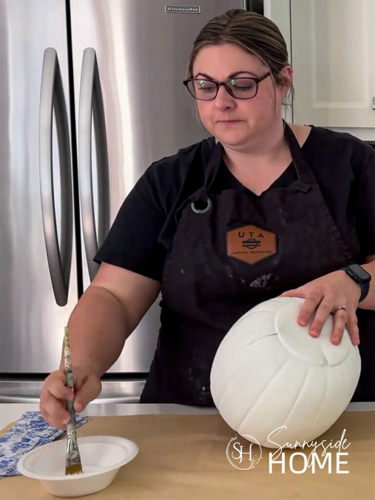 Woman applies Mod Podge decoupage medium to a section of a white painted pumpkin.