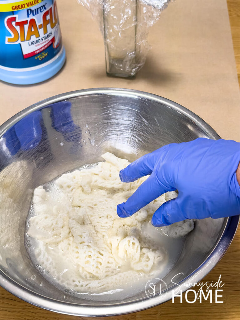 A woman wearing blue latex gloves places a vintage doily in a stainless steel bowl filled with fabric starch.