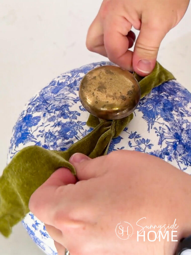 Woman ties a green velvet ribbon around the brass knob stem on a chinoiserie pumpkin.