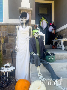 Fun outdoor Halloween decorations: a pair of skeletons dressed up as Frankenstein and the bride of Frankie with thrift store clothes, wig, and hat. In the background the front door is decorated with a ghost wreath and a skeleton dressed up as Count Dracula.