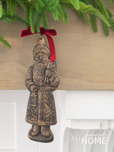 Finished faux aged brass Santa ornaments embellished with a red velvet bow are hung from green garland on the mantle.