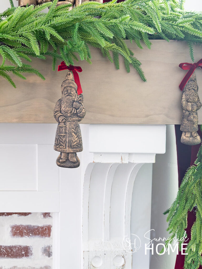 Finished faux aged brass Santa ornaments embellished with a red velvet bow are hung from green garland on the mantle.