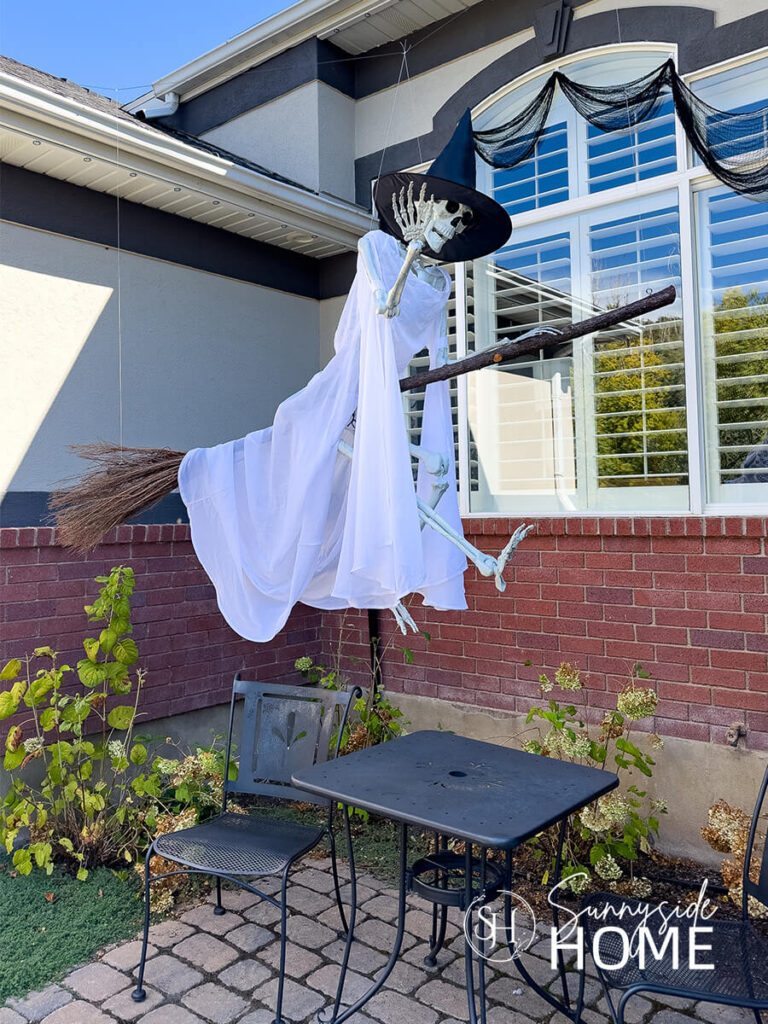 A skeleton is dressed for Halloween in a white flowing thrift store dress and witch's hat, flying through the air on her broom.